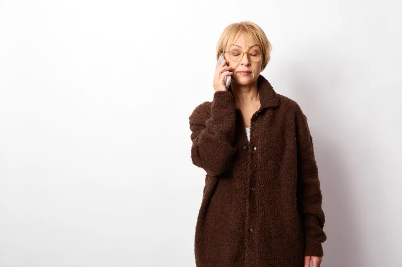 An elderly woman with glasses and a brown coat holds a phone to her ear, looking tired and stressed, pausing in studio isolation on a white background, communication concept