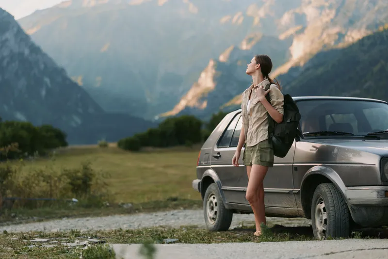 Young woman traveler with backpack stands near car on mountain road looks at scenic landscape enjoys nature and outdoor adventure lifestyle in summer day.