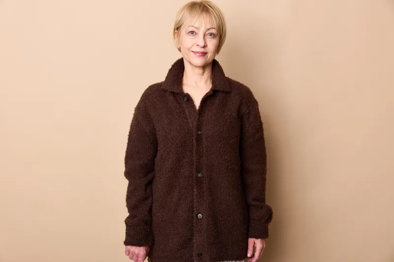 Woman senior portrait coat standing in a studio, beige background, calm mature expression, wearing dark fuzzy coat, simple fashion concept about aging and self confidence
