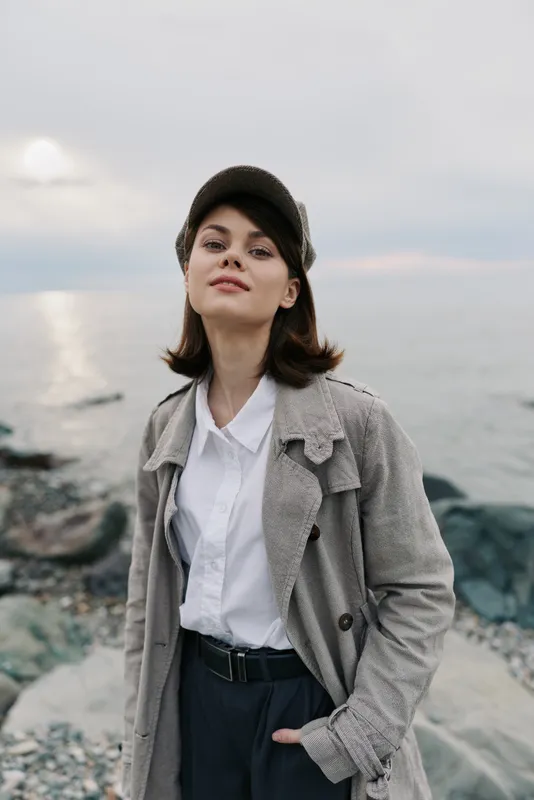 Young woman wearing a textured coat and cap stands confidently near rocky shore with ocean and cloudy sky background. Casual fashion and outdoor lifestyle concept.