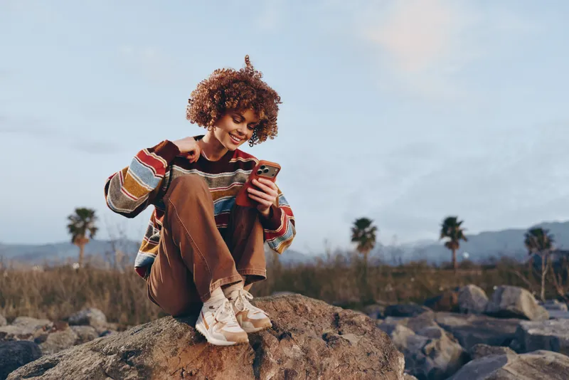 Woman lifestyle outdoors, smiling on a rock, holding cellphone and posing for selfie, curly hair and casual sweater, scenic background with palm trees under cloudy sky, travel concept