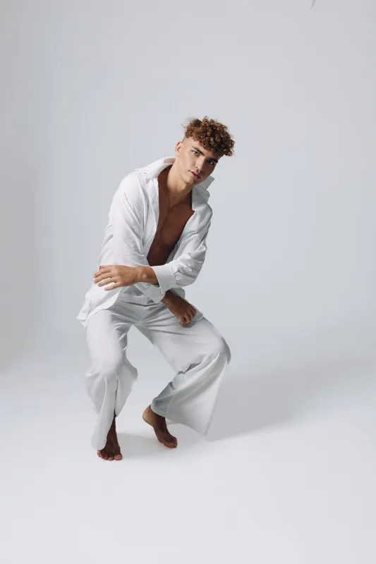 Young man with curly hair wearing white loose clothing posing barefoot in studio with neutral background for fashion, style, and modern casual outfit concept.
