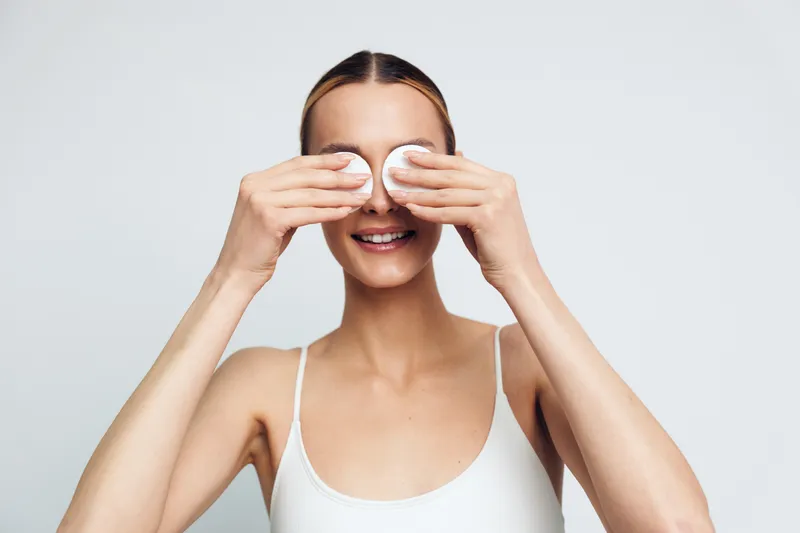 Young woman with blonde hair in a white tank top covers her eyes with cotton pads while smiling against a plain light background in a skincare routine moment.