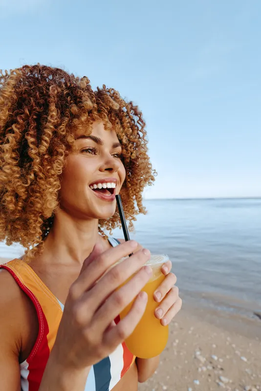Young woman with curly hair on a sunny beach enjoys a glass of orange juice with a straw, smiling as the blue sea and clear sky frame her in bright summer light.
