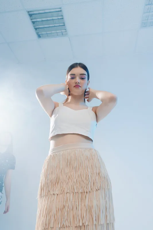Young woman beauty fashion model poses with hands behind head wearing fringed skirt and white top in bright studio. Concept for style, elegance, and modern trends.