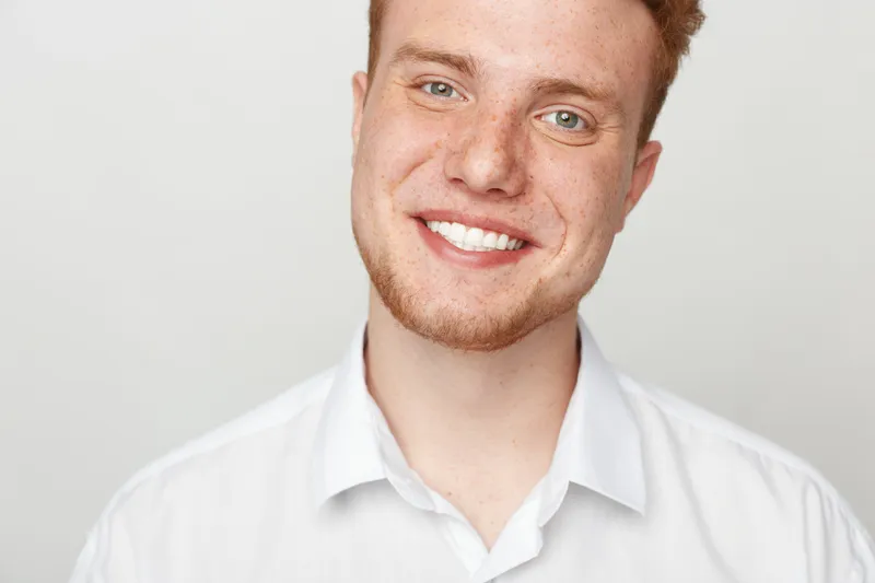 Smiling young man with red hair and freckles wears white shirt, looking at camera on light background. Concept of happiness, confidence, youth, and natural beauty in portrait photography.