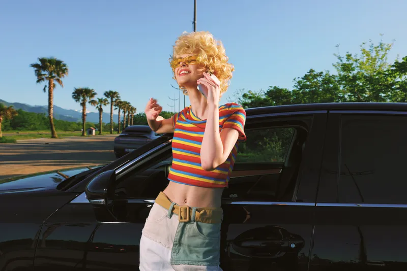 Woman with curly orange wig in a striped crop top leans against a dark car in a sunny outdoor scene, retro film color, casual fashion, playful mood, summer vibe.