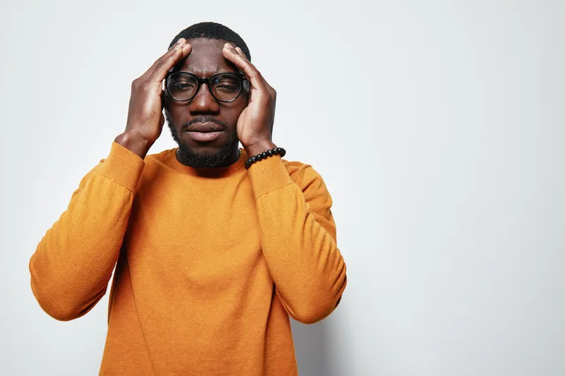 African man with dark skin and glasses wearing orange sweater holding head with hands, expressing stress or headache on white background. Male adult, casual clothing and emotional discomfort.