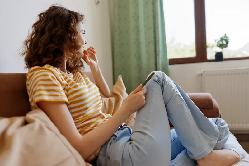 Young woman with curly brown hair sits on a sofa in a bright home, wearing a striped yellow tee and jeans, calmly scrolling a smartphone with a thoughtful mood.