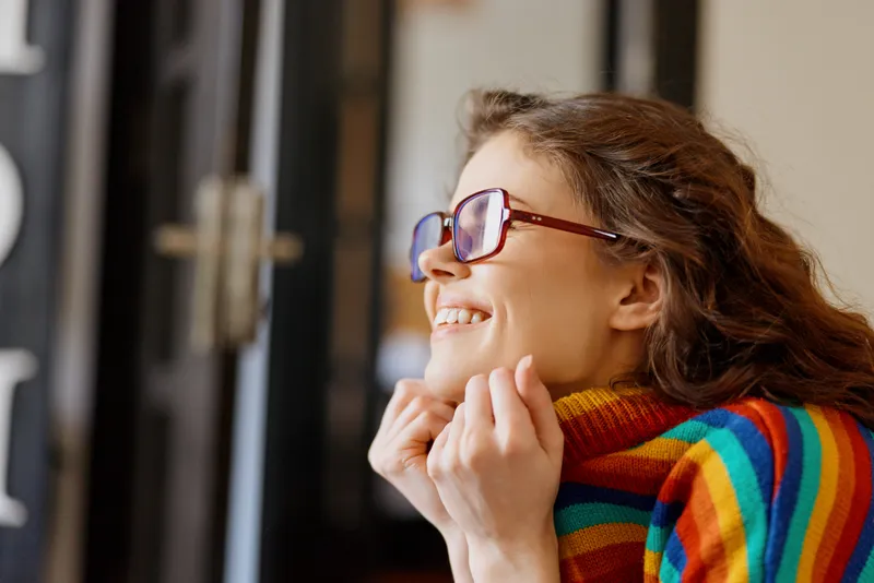 Cheerful woman indoors in a colorful striped scarf and glasses, smiling as she looks out the window, enjoying a cozy home lifestyle moment with warm natural light.