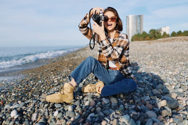 Beach photographer woman with camera captures waves on a rocky shoreline; casual plaid shirt, jeans and boots highlight an outdoor moment in bright sunlight and sea breeze.