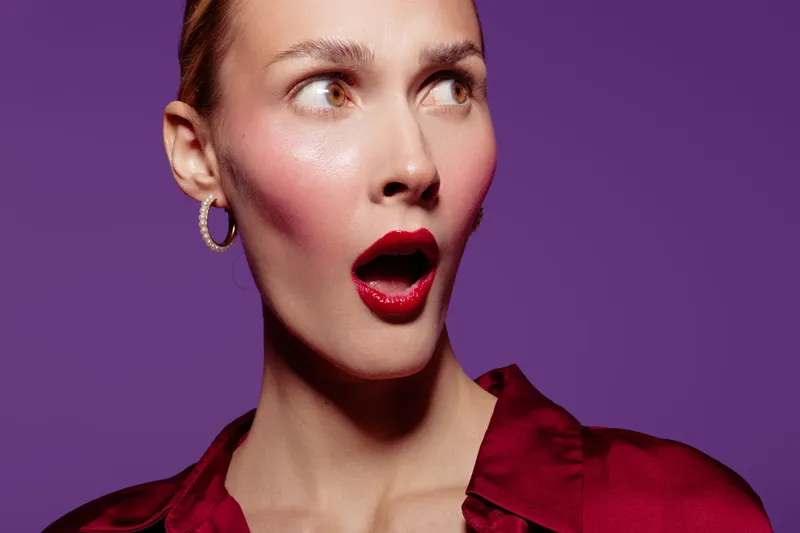 Surprised woman with red lipstick and earrings looks sideways on purple background. Close-up portrait with expressive face and shiny skin. Concept for emotions, beauty, and fashion.