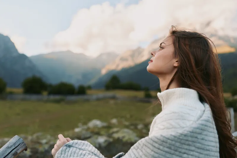 Young woman with long brown hair wrapped in a cozy gray blanket enjoys a peaceful moment outdoors with mountains and cloudy sky in the background.