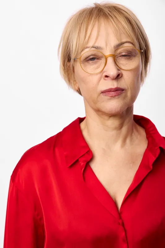 Senior woman portrait, short blonde hair, round glasses, red blouse in a clean studio background, thoughtful calm expression, highlighting aging, confidence, wellbeing and selfcare