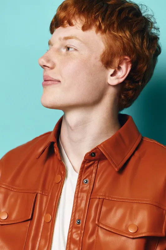 Redhead man with curly hair wearing a brown leather jacket and white shirt looking sideways against turquoise background in studio portrait.