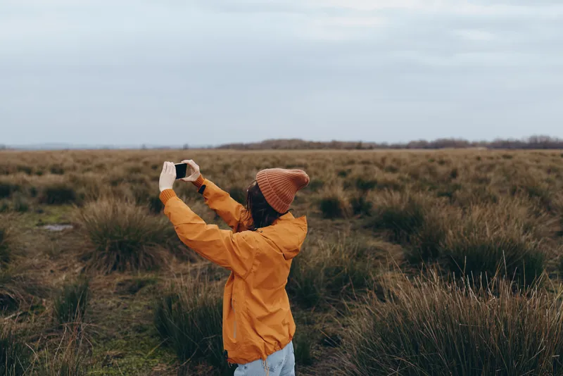 Woman outdoors in a meadow of grassland, wearing an orange jacket and knitted hat, lifting a phone to capture nature, peaceful lifestyle, soft daylight, muted colors, windy countryside landscape