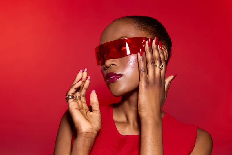 Stylish African American woman with dark skin wearing red futuristic glasses poses against red background. Concept of fashion, beauty, modern style, and confident female model in studio.