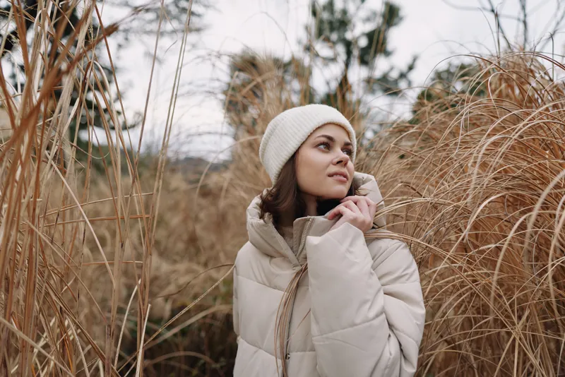 Woman in a light puffer coat and white beanie stands among tall grasses, outdoors in muted winter light, thoughtful pose, soft textures, natural backdrop, seasonal fashion portrait, showcasing quiet