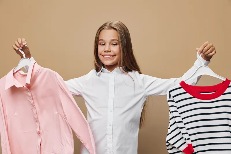Girl with long hair smiling and holding two clothes hangers with different shirts indoors. Child in white blouse choosing outfit for casual or formal wear.