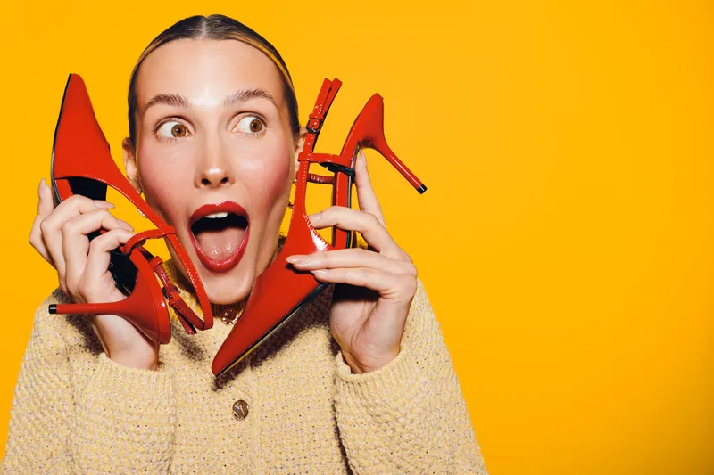 Excited woman holds red high heel shoes near face on yellow background. Concept of fashion, shopping, style, and vibrant emotions with bright studio lighting and casual outfit.