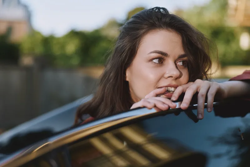 Brunette woman leans through a car window and bites her lip in a candid moment that hints at flirtation and spontaneity, with soft natural light highlighting features and a glossy vehicle