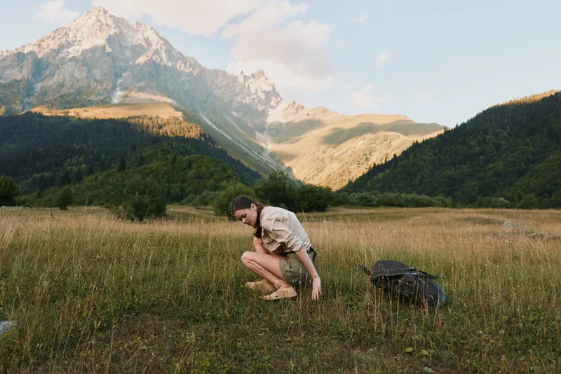 Woman in outdoor meadow with distant mountains, crouching amid grass, backpack nearby, embracing sunlight and travel vibe, natural portrait of exploration