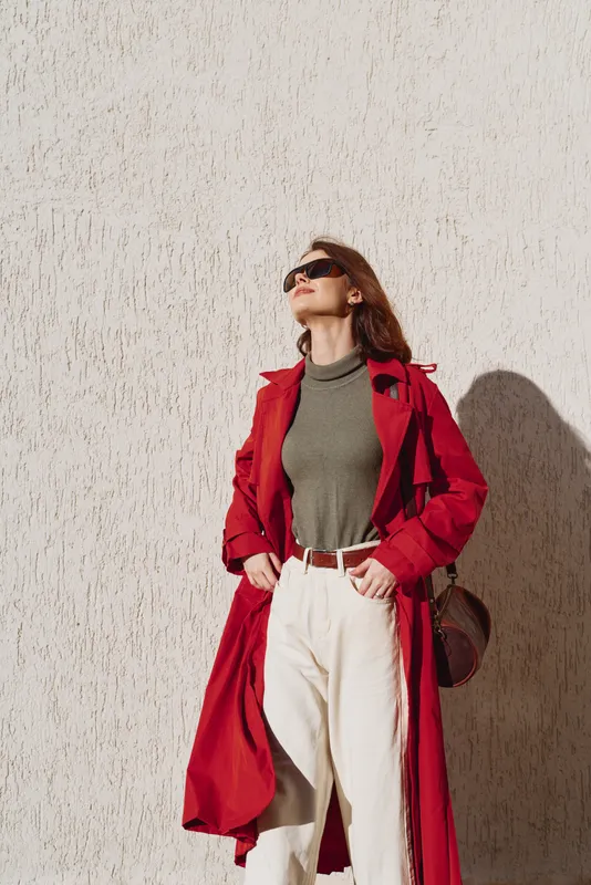 A confident woman in a red trench coat and sunglasses stands against a textured white wall looking upwards. Casual outfit with high waist pants and a brown bag.
