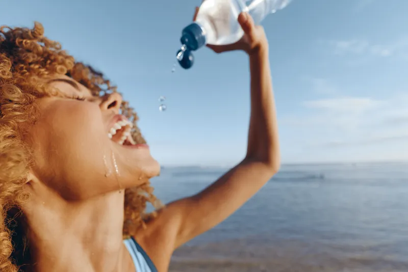 Young woman with curly hair tilts her head back, spraying water from a bottle at the sunny seaside, capturing a refreshing, carefree moment and bright mood.