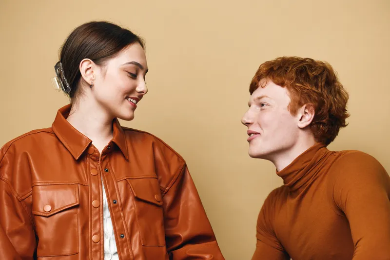Young man and woman smiling and looking at each other for friendship and positive emotion on beige background. Casual style with red hair and brown leather jacket in studio.