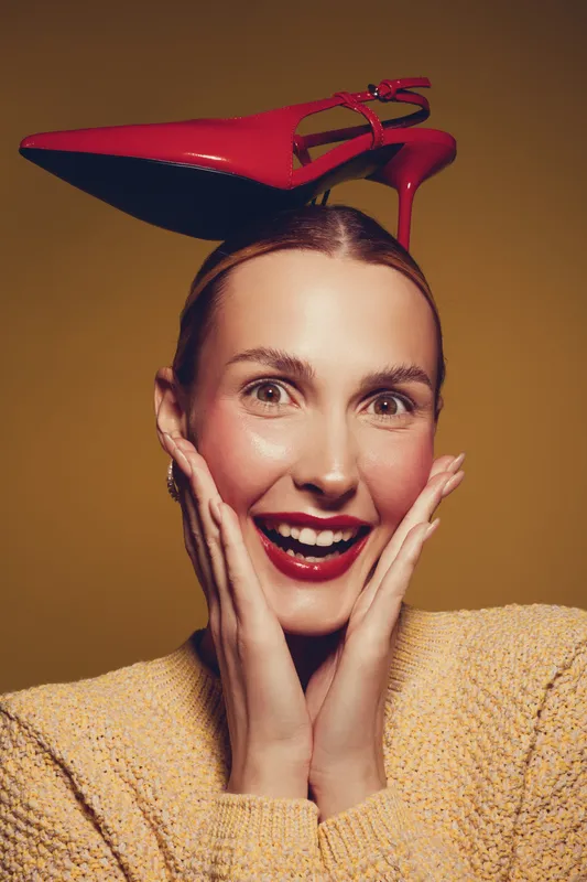 Smiling woman with red high heel shoe on head expresses joy and surprise, wearing yellow sweater on brown background. Concept of fashion, fun, and playful style.