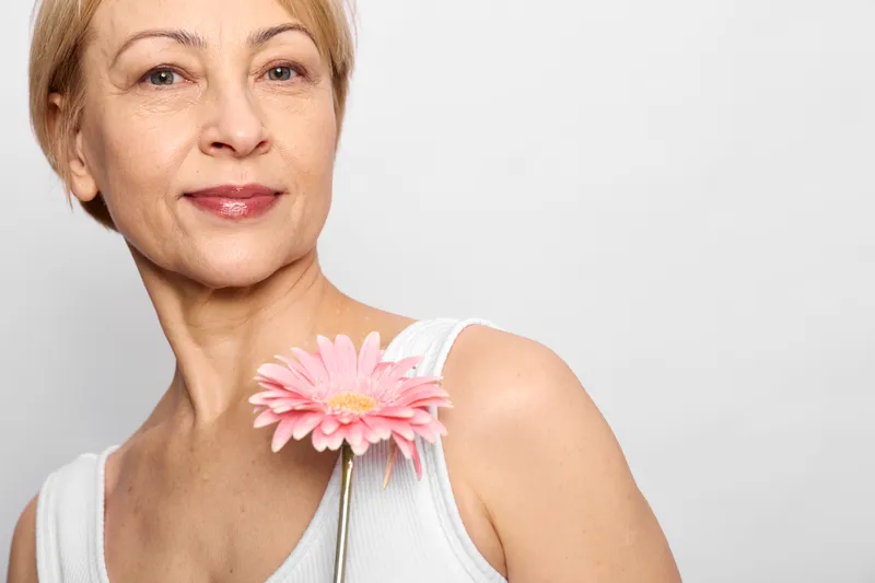 Mature woman, skincare, selfcare, flower, studio portrait, smiling softly with a pink gerbera on white background, soft lighting shows healthy skin, wellness and calm mood