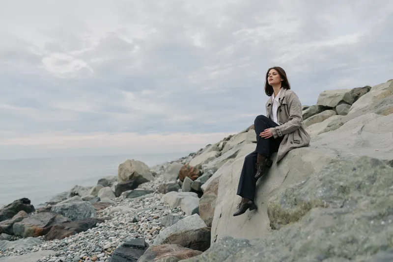 A calm woman in casual clothes sits on large rocks near the sea under a cloudy sky. The peaceful coastal scene conveys relaxation and nature connection.