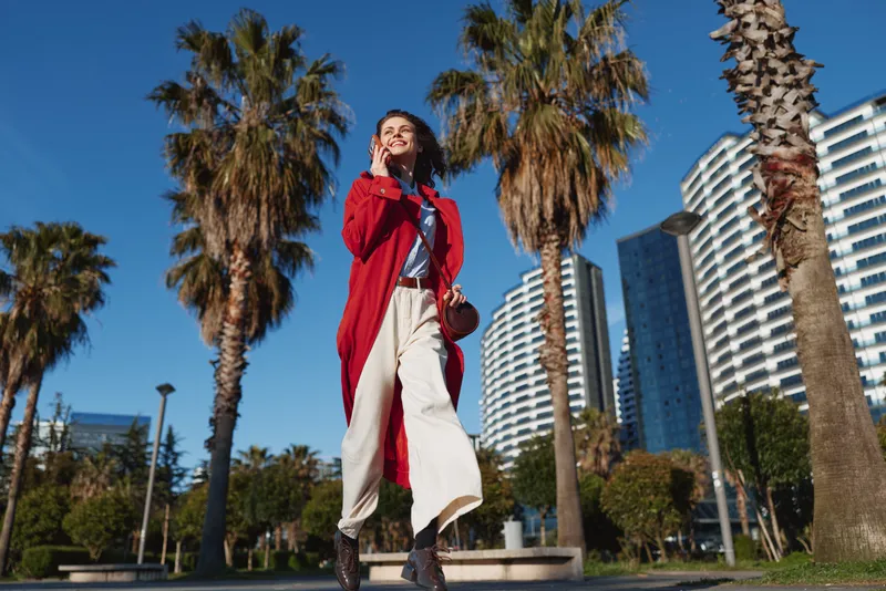 Young woman in a red coat walks confidently on a sunny urban street with tall palm trees and modern buildings behind, exuding energy and carefree confidence.
