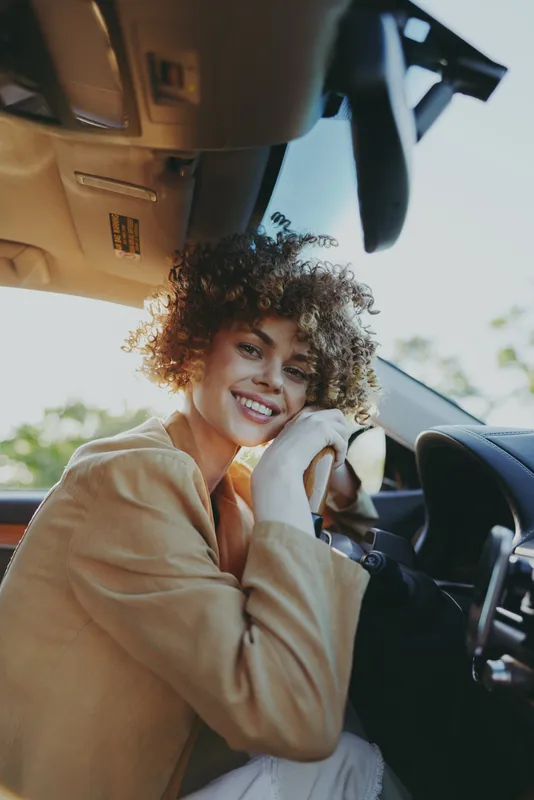 Cheerful woman sits behind the steering wheel, smiling at the camera in a sunlit car interior. Warm retro color tones create a casual lifestyle moment.