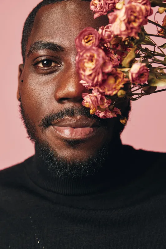 African man with dark skin and beard wearing black turtleneck posing with dried pink flowers on face in creative conceptual visual style with soft pink background.