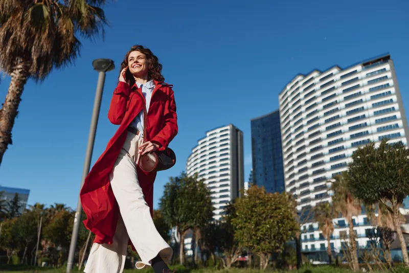 Young woman in a red coat walks confidently through a sunny urban park with modern buildings and palm trees in the background.