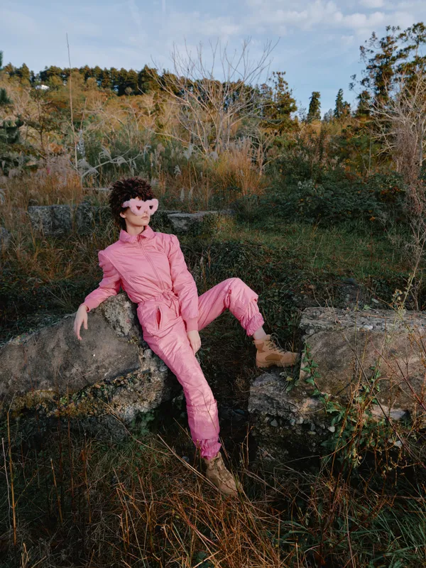 Outdoor model in a pink jumpsuit lounges on rock ruins, blending fashion and nature with a relaxed pose, bold color, textured light and atmospheric mood.