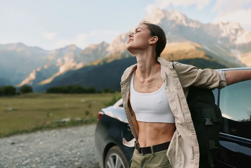 Young woman with brunette hair stretches beside a parked car in a vast mountain landscape, feeling free and energized as the open space surrounds her, signaling adventure and wellness.