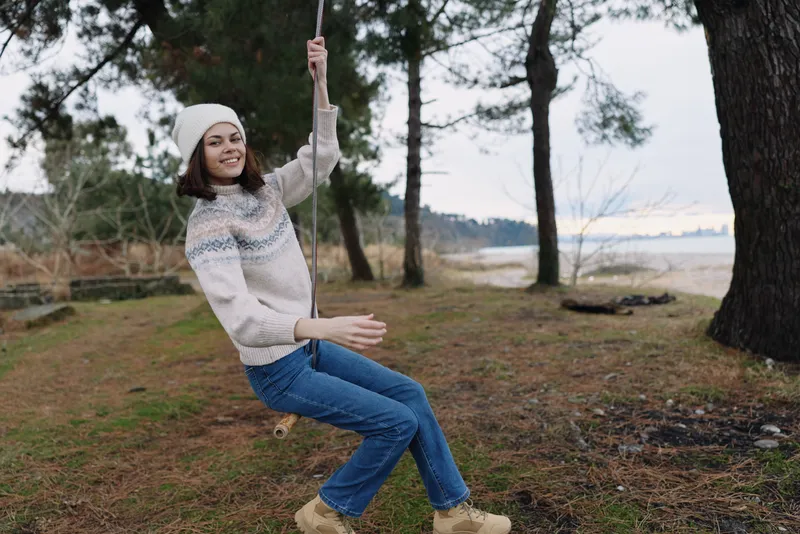 Outdoor swing ride with a smiling girl in a cozy knit sweater and beanie by a lakeside forest, capturing playful carefree mood and seasonal leisure in nature.