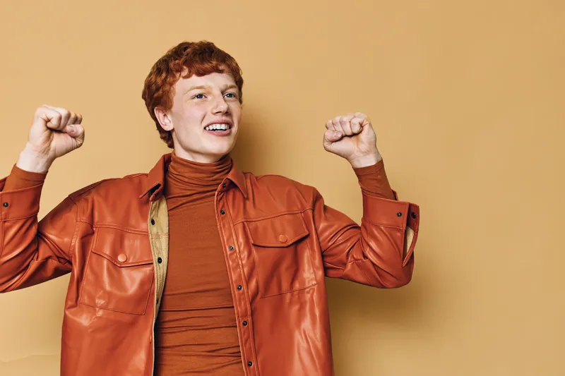 Young man with red hair wearing orange jacket and turtleneck, smiling and raising fists with excitement against plain beige background. Casual confident happy emotion.