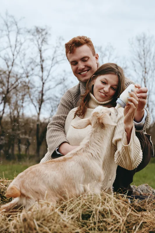 A young couple feeds a baby goat with a bottle while sitting on hay in an outdoor natural setting during autumn, enjoying a relaxed lifestyle moment together.