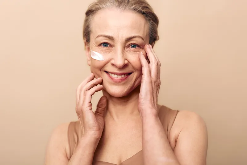 Smiling mature woman with light skin applies white face cream on cheek, touching face gently. Beige background and natural light create calm mood. Concept of skincare, aging, and beauty care.