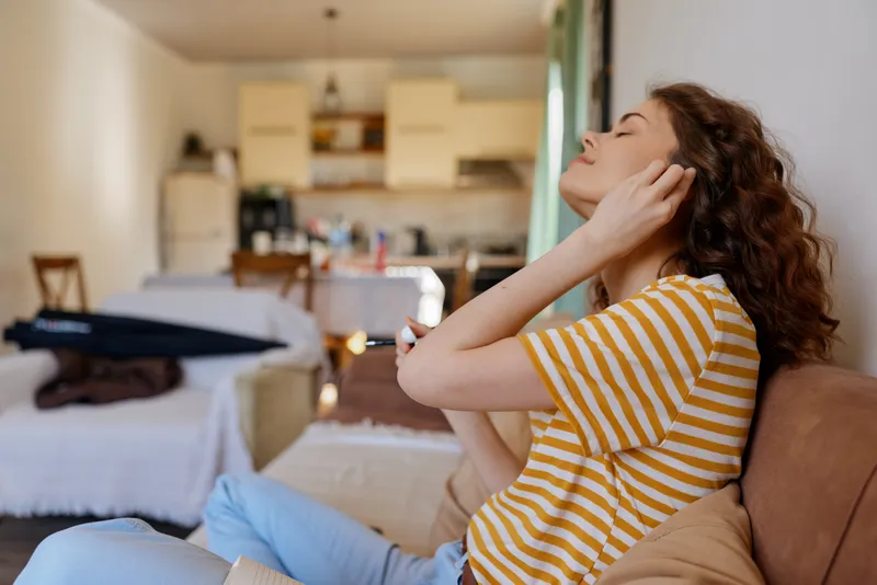 Woman with striped yellow shirt relaxing at home, eyes closed, listening to music. Cozy living space with soft lighting and warm tones creating a peaceful mood. Comfortable cushions, wooden
