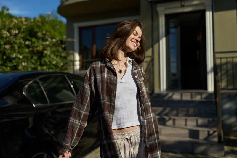 Portrait of a smiling woman enjoying a sunny day outside a modern home, casual lifestyle image, plaid shirt over a white top, relaxed mood, candid friendly moment