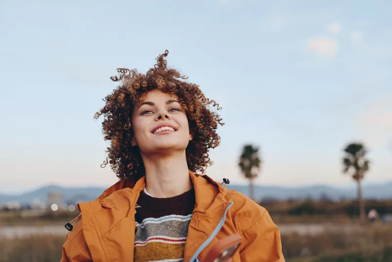 Cheerful woman outdoors, smiling upward with curly hair in wind, warm jacket and striped sweater, sunlit sky, relaxed lifestyle moment, soft breeze, open field background