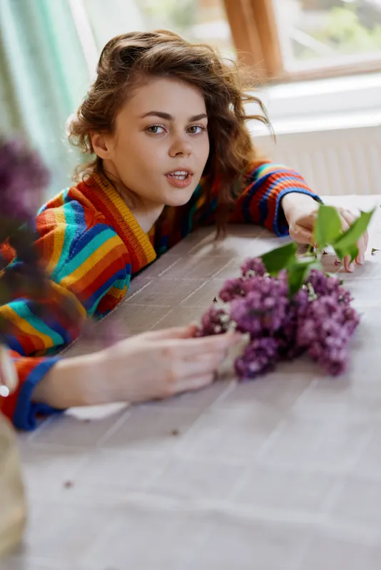 Cheerful woman indoors, relaxed lifestyle moment, colorful sweater, soft natural light, flowers on table, cozy home atmosphere, candid mood, warm colors, casual pose, everyday comfort, homely setting