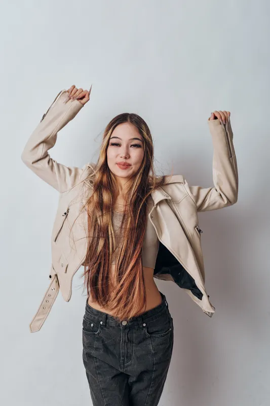 Young woman with long hair wearing beige jacket and black jeans posing with raised arms against plain background, expressing confidence and casual style fashion.