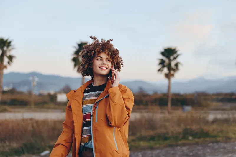 Woman lifestyle portrait outdoors, curly hair, orange jacket, holding phone near ear, smiling and speaking, countryside road with palm trees and distant mountains, sunny travel moment