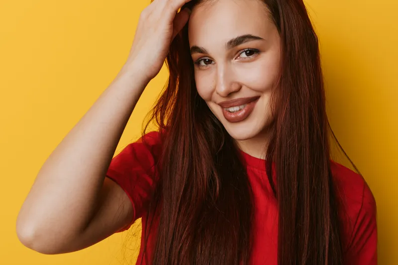 Young woman with long brown hair smiling and looking at camera on yellow background. Casual style with red shirt and natural makeup for portrait photo.