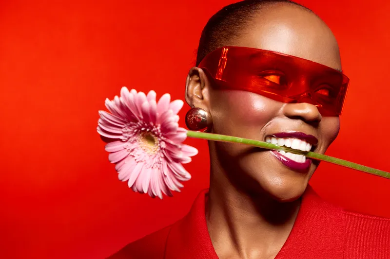 Portrait of a smiling black woman with dark skin wearing red glasses and a red outfit holding a pink flower in her mouth on a red background. Concept of joy and fashion.